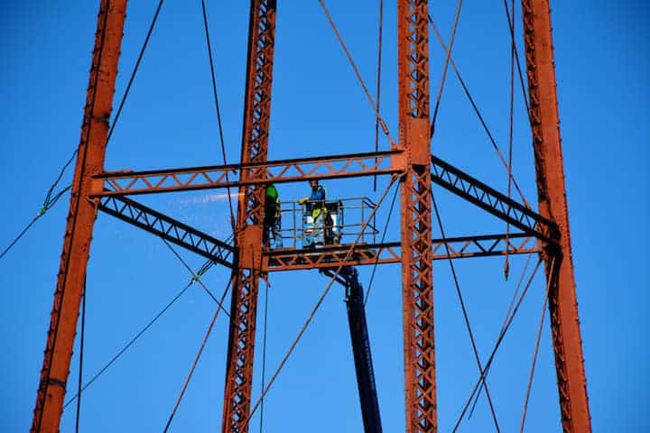 Two men in a bucket lift cut several supports and girders on the tower, weakening it enough to allow a backhoe to pull it over with cables. 