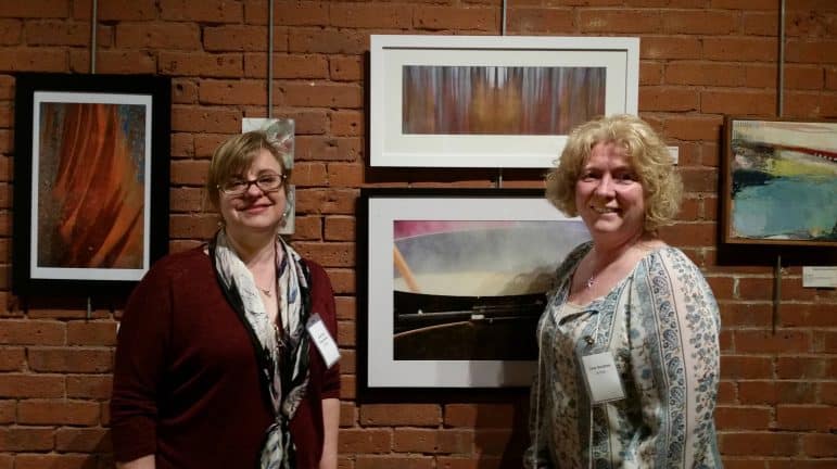 [CREDIT: Mary Carlos] Exhibiting artists Katerina Stepanova (her piece is to the left of her “The Boat Rust) and Lisa Bushee (her piece is above her head “Fall Pano”) stand with their artwork at Warwick Center for the Arts. 