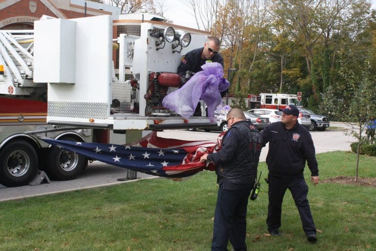 [CREDIT: Beth Hurd] EGFD Firefighters Kevin King and Dan Andrade, joined by Lt. Stephen Babcock, fasten a purple bow and prepare to hang the American Flag from the basket of their Ladder 1 on Tuesday, Nov. 7, the truck parked in front of the East Greenwich Police Station.