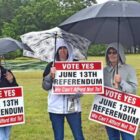[CREDIT: Robert Ford] Three English teachers from Coventry High School try and stay dry as they stand at the entrance to Westwood Estates polling station urging those casting ballots in the June 13 budget election to vote “Yes.”