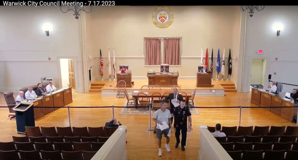 [CREDIT: City of Warwick] A Warwick Officer escorts Rob Cote from Council Chambers July 17, 2023. Councilwoman Donna Travis ordered him removed for persisting in speaking about a property dispute detailed in a Providence Journal article involving Travis.